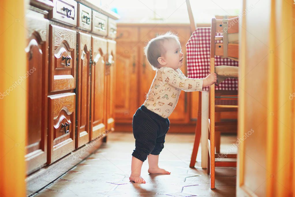 Baby girl standing on the floor in the kitchen and holding on to furniture. Little child oulling up at home