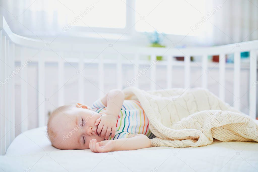 Adorable baby girl sleeping in co-sleeper crib attached to parents' bed. Little child having a day nap in cot. Sleep training concept. Infant kid in sunny nursery