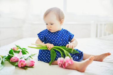 Mavi elbiseli bebek bir demet pembe laleyle oynuyor. Güneşli yuvada küçük bir çocuk. Bebek çiçeklerle eğleniyor.