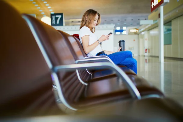 国际机场的年轻女子带着行李和咖啡要走 等着航班 看着手机 — 图库照片