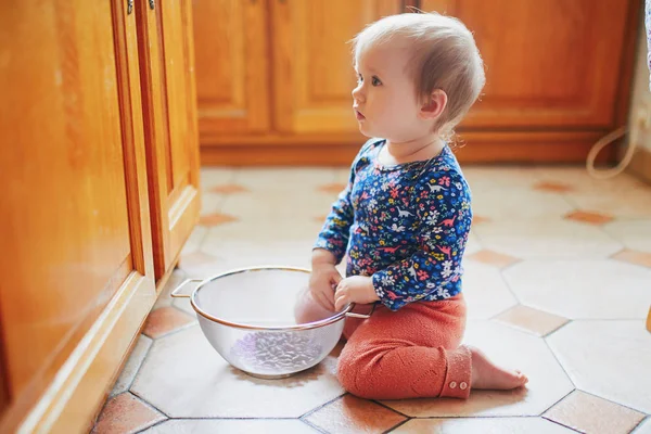 Niña sentada en el suelo en la cocina y jugando con utensilios de cocina — Foto de Stock