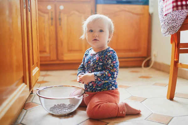 Niña sentada en el suelo en la cocina y jugando con utensilios de cocina —  Fotos de Stock