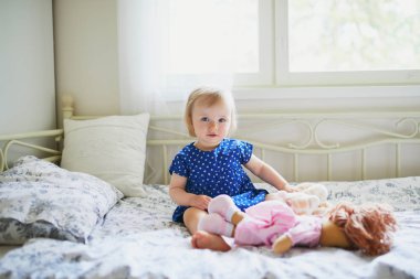 Mavi elbiseli sevimli kız yatakta oturan ve bir bebek ile oynarken