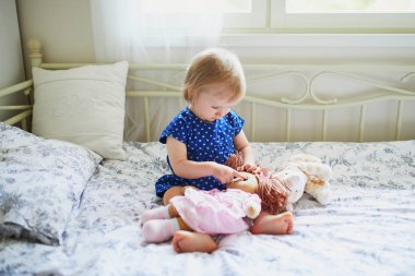 Mavi elbiseli sevimli kız yatakta oturan ve bir bebek ile oynarken