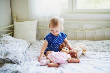 Mavi elbiseli sevimli kız yatakta oturan ve bir bebek ile oynarken