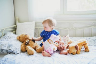 Mavi elbiseli sevimli kız yatakta oturan ve doldurulmuş oyuncaklar ile oynarken