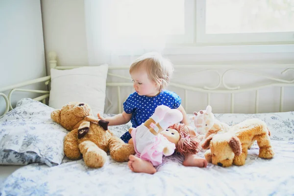Очаровательная девочка в синем платье сидит на кровати и играет с игрушками — стоковое фото