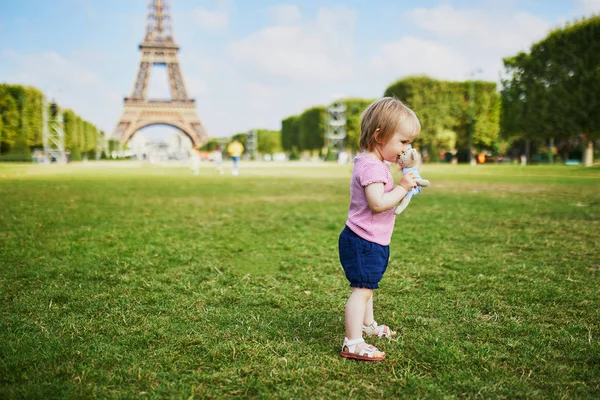 Happy toddler girl with her favorite teddy bear standing on the grass near the Eiffel tower in Paris, France — Stock Photo, Image
