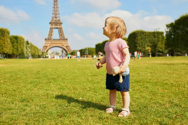 Happy toddler girl with her favorite teddy bear standing on the grass near the Eiffel tower in Paris, France — Stock Photo, Image