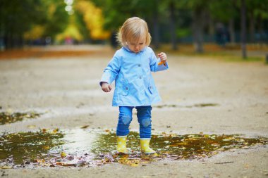 Sarı çizme giyen ve sonbahar günü su birikintisine atlayan bir çocuk. Tatlı küçük kız yağmurlu bir günde parkta su ve çamurla eğleniyor. Çocuklar için açık hava sonbahar aktiviteleri