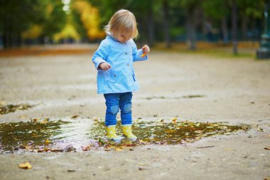 Sarı çizme giyen ve sonbahar günü su birikintisine atlayan bir çocuk. Tatlı küçük kız yağmurlu bir günde parkta su ve çamurla eğleniyor. Çocuklar için açık hava sonbahar aktiviteleri
