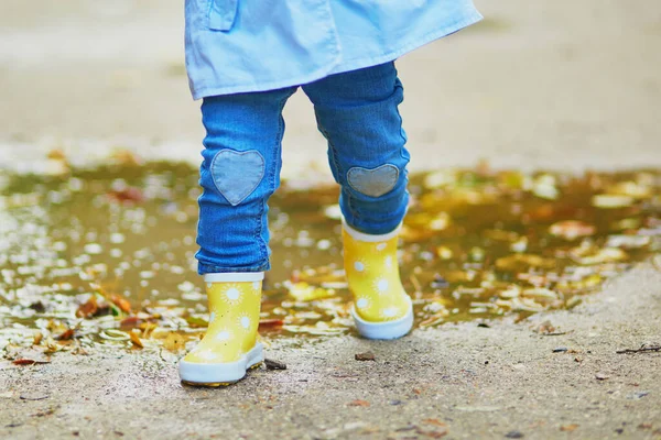 Child wearing yellow rain boots and jumping in puddle on a fall day. Toddler girl having fun with water and mud in park on a rainy day. Outdoor autumn activities for kids