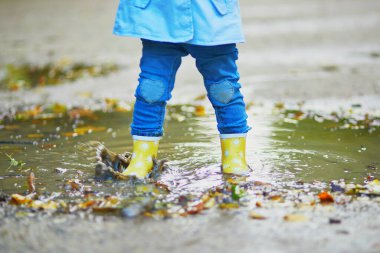 Sarı çizme giyen ve sonbahar günü su birikintisine atlayan bir çocuk. Küçük kız yağmurlu bir günde parkta su ve çamurla eğleniyor. Çocuklar için açık hava sonbahar aktiviteleri