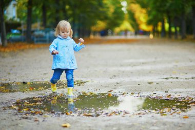 Sarı çizme giyen ve sonbahar günü su birikintisine atlayan bir çocuk. Tatlı küçük kız yağmurlu bir günde parkta su ve çamurla eğleniyor. Çocuklar için açık hava sonbahar aktiviteleri
