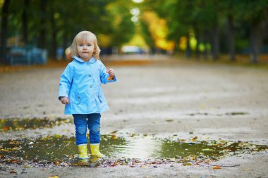 Sarı çizme giyen ve sonbahar günü su birikintisine atlayan bir çocuk. Tatlı küçük kız yağmurlu bir günde parkta su ve çamurla eğleniyor. Çocuklar için açık hava sonbahar aktiviteleri