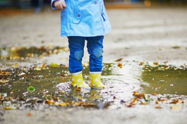 Sarı çizme giyen ve sonbahar günü su birikintisine atlayan bir çocuk. Küçük kız yağmurlu bir günde parkta su ve çamurla eğleniyor. Çocuklar için açık hava sonbahar aktiviteleri