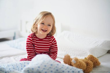 Çizgili kırmızı ve beyaz pijamalı mutlu küçük kız uyandıktan hemen sonra yatakta oturuyor. Küçük çocuklar için gündüz şekerlemeleri