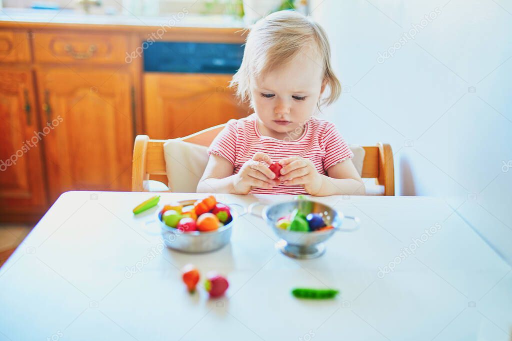 Adorable little girl playing with toy fruits and vegetables at home, in kindergaten or preschool. Indoor creative games for kids