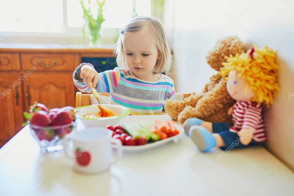 Adorable toddler girl eating fresh fruits and vegetables for lunch. Child feeding doll and teddy bear in the kitchen. Delicious healthy food for kids