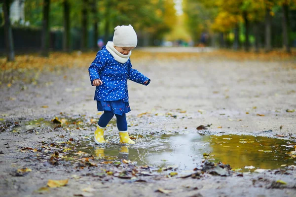 Child wearing yellow rain boots and jumping in puddle on a fall day. Adorable toddler girl having fun with water and mud in park on a rainy day. Outdoor autumn activities for kids