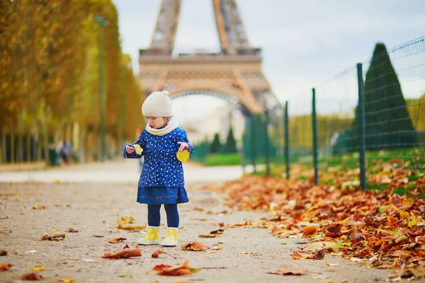 青いレインコートを着た愛らしい幼児の女の子は フランスのパリのエッフェル塔の近くに黄色の落ち葉を集めています 暖かく晴れた秋の日を楽しむ幸せな子供 — ストック写真