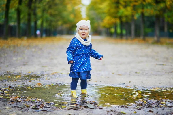 Child wearing yellow rain boots and jumping in puddle on a fall day. Adorable toddler girl having fun with water and mud in park on a rainy day. Outdoor autumn activities for kids