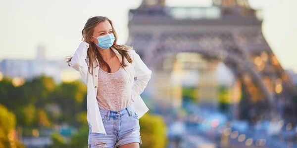 Young girl standing near the Eiffel tower in Paris and wearing protective face mask during coronavirus outbreak. Pandemic and lockdown in France. Tourist spending vacation in France after quarantine