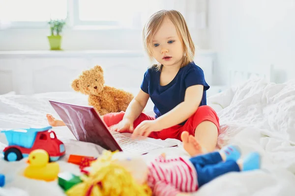 Toddler girl with laptop and toys in bed. Kid using gadget to communicate with friends or kindergartners. Education, distance learning or work from home with kids. Stay at home entertainment