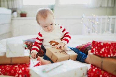Sıcak bir Noel süveteri giyip Noel hediyelerini ilk Noel 'inde açan mutlu küçük bir kız bebek. Evde çocuklarla Noel 'i kutluyoruz.