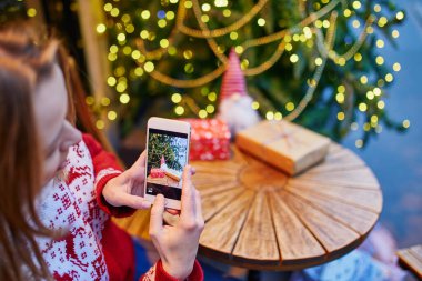 Kahve fincanının ve Noel hediyesinin mevsimsel tatiller için dekore edilmiş kafede fotoğrafını çeken bir kadın.