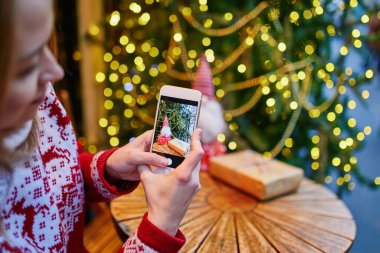 Kahve fincanının ve Noel hediyesinin mevsimsel tatiller için dekore edilmiş kafede fotoğrafını çeken bir kadın.