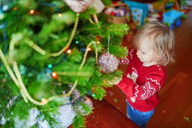 Noel ağacı süveteri giyip Noel ağacı süsleyen sevimli küçük bir kız. Evde çocuklarla mevsimlik tatilleri kutluyoruz.