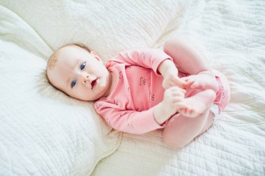 Bebek kız yatakta sırt üstü yatar ve ayak parmaklarına dokunur. Pembe elbiseli sağlıklı küçük bir çocuk. Güneşli kreşte küçük bir çocuk.
