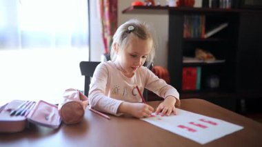 7 yaşında bir kız, evde bir masada oturuyor ve ödevini konsantre bir şekilde yapıyor. Okul malzemeleriyle çevrili, kağıt üzerine kalemle yazıyor.