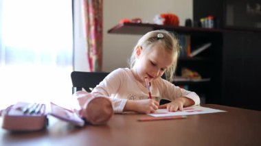 7 yaşında bir kız, evde bir masada oturuyor ve ödevini konsantre bir şekilde yapıyor. Okul malzemeleriyle çevrili, kağıt üzerine kalemle yazıyor.