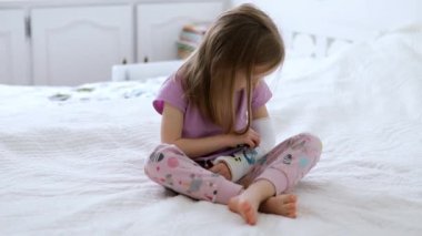 Adorable preschooler girl with a broken arm at home on the bed draws with felt-tip pens on an orthopedic cast. How to have fun with a broken limb. Broken arm in a cast in kids