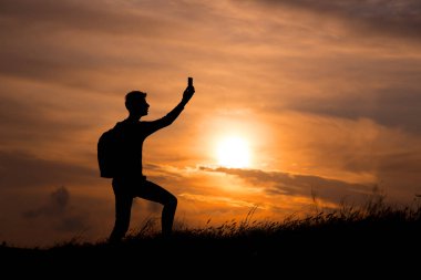 Turist silueti bir dağın tepesine uzanmış günbatımının tadını çıkarıyordu. Spor ve aktif yaşam konsepti. Adam cep telefonuyla gün batımının fotoğraflarını çekiyor.