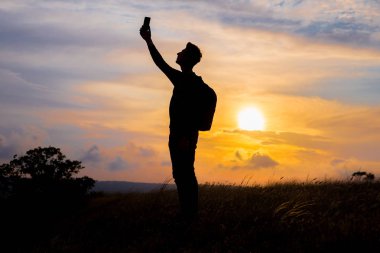 Turist silueti bir dağın tepesine uzanmış günbatımının tadını çıkarıyordu. Spor ve aktif yaşam konsepti. Adam cep telefonuyla gün batımının fotoğraflarını çekiyor.