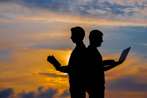 silhouette of a men with laptop and a book on sunset or sunrise background. The idea of the contradictions of the old and new values. The concept of old and modern