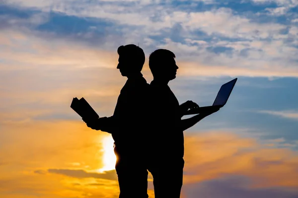 silhouette of a men with laptop and a book on sunset or sunrise background. The idea of the contradictions of the old and new values. The concept of old and modern