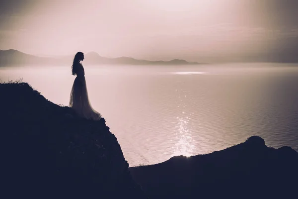 穿着长裙的年轻女子站在岩石上看着大海 — 图库照片