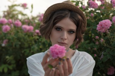 Bahçede güllerin yanında poz veren kıvırcık saçlı güzel bir genç kadın. Parfüm reklamı konsepti.