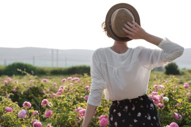 Bahçede güllerin yanında poz veren kıvırcık saçlı güzel bir genç kadın. Parfüm reklamı konsepti.