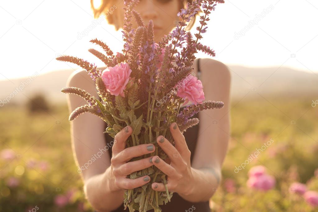 Beautiful young woman with curly hair posing near roses in a garden. The concept of perfume advertising.