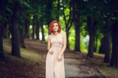 Pembe elbise ve Kızıl saçlı, güzel genç kadın için bir tarih bekliyor. Yaz şehir, pembe çiçekli çalılar adımlarında bir gezinti