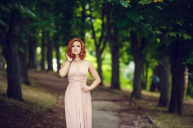 Pembe elbise ve Kızıl saçlı, güzel genç kadın için bir tarih bekliyor. Yaz şehir, pembe çiçekli çalılar adımlarında bir gezinti