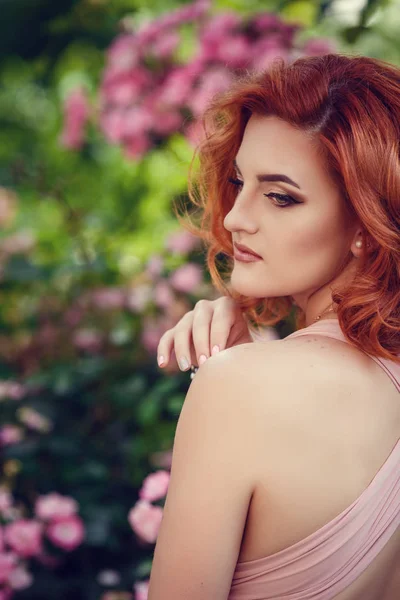 Pembe elbise ve Kızıl saçlı, güzel genç kadın için bir tarih bekliyor. Yaz şehir, pembe çiçekli çalılar adımlarında bir gezinti