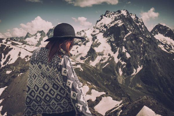 Boho woman wearing hat and poncho standing by the mountain. Cold weather, snow on hills. Winter hiking. Wanderlust.