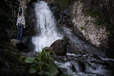 Waterfall tarafından ayakta ve bakarak panço ve şapka giyen Boho kadın.
