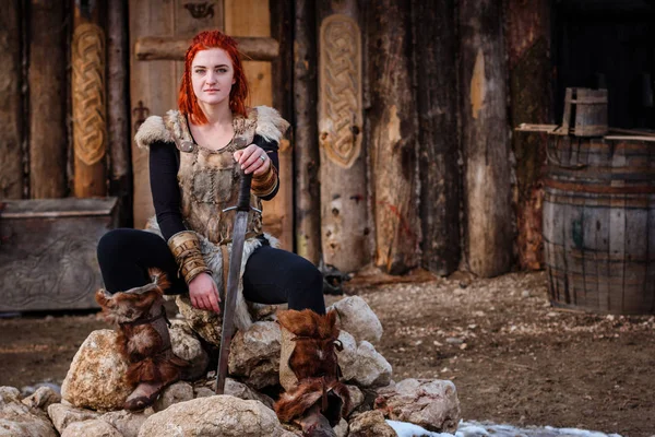 Red-haired woman is a Viking. Against the backdrop of a village or an ancient settlement. In the hands of a sword — Stock Photo, Image